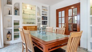 A dining room with a glass-top table surrounded by wooden chairs, white shelves with various items, and a wooden door with glass panels. - Holiday Home 'Villa Jordi Y Nuri' with Private Terrace, Wi-Fi and Air Conditioning (Cambrils)