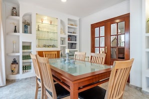 Une salle à manger avec une table à plateau de verre entourée de chaises en bois, des étagères blanches où sont disposés divers objets, et une porte en bois dotée de panneaux de verre.