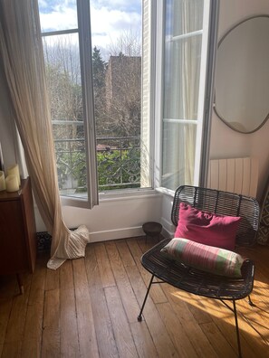 Une pièce avec une grande fenêtre, un fauteuil en osier noir agrémenté d’un coussin rose et un radiateur blanc.