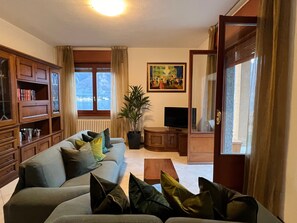 Ein Wohnzimmer mit einer Couch, einem Fernseher, einem Bücherregal und einem Fenster mit Vorhängen.