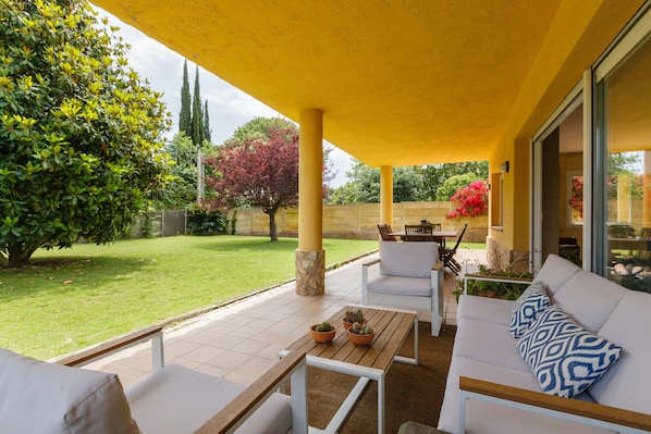 An outdoor patio area with a yellow roof, a white sofa with patterned cushions, a wooden table, and a potted plant.
