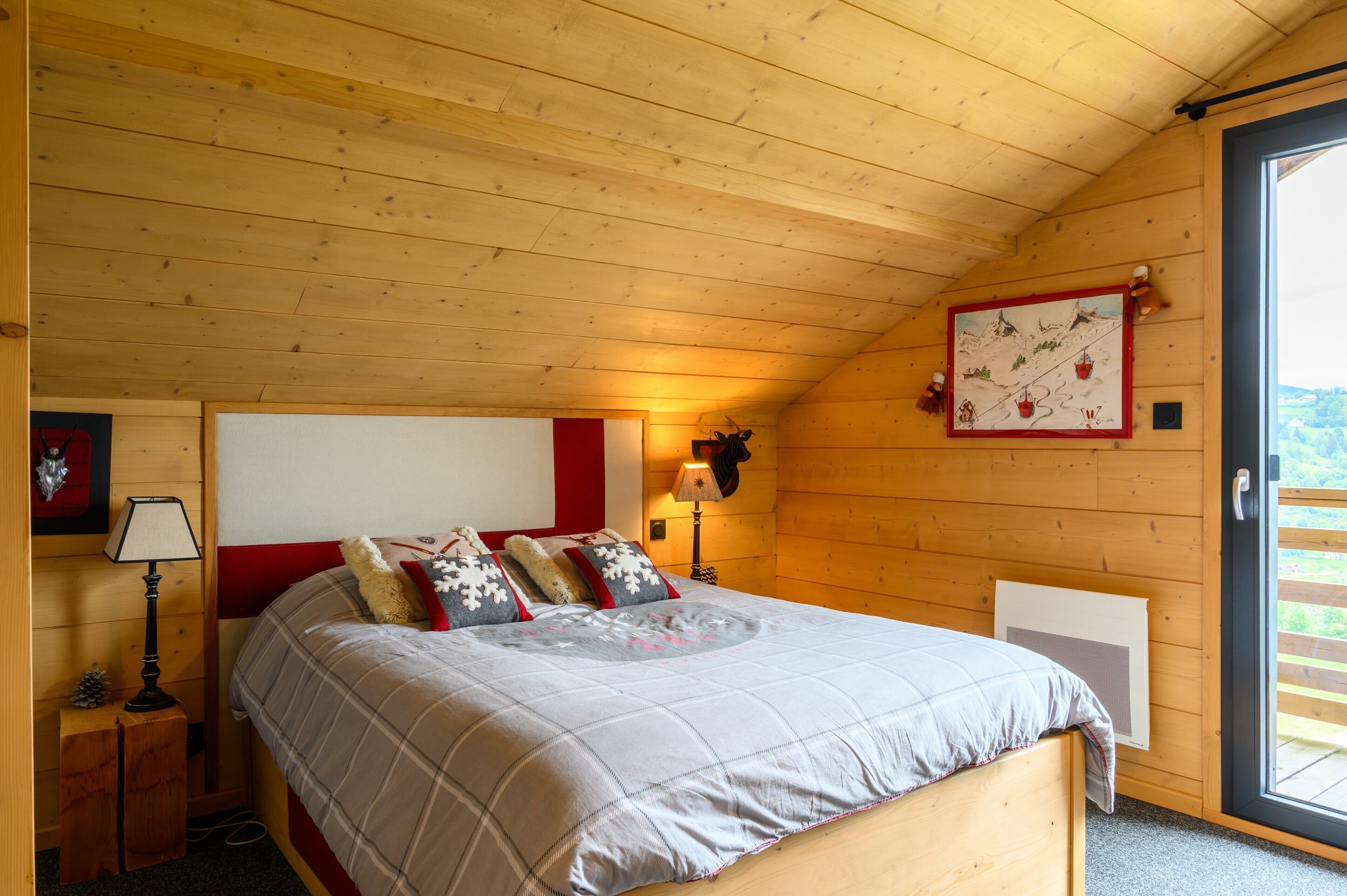 A wooden cabin bedroom with a bed, a nightstand, a lamp, a wall-mounted heater, and a window with a view.