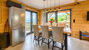 A modern kitchen with a refrigerator, dining table with chairs, and large windows offering a view of the outdoors.