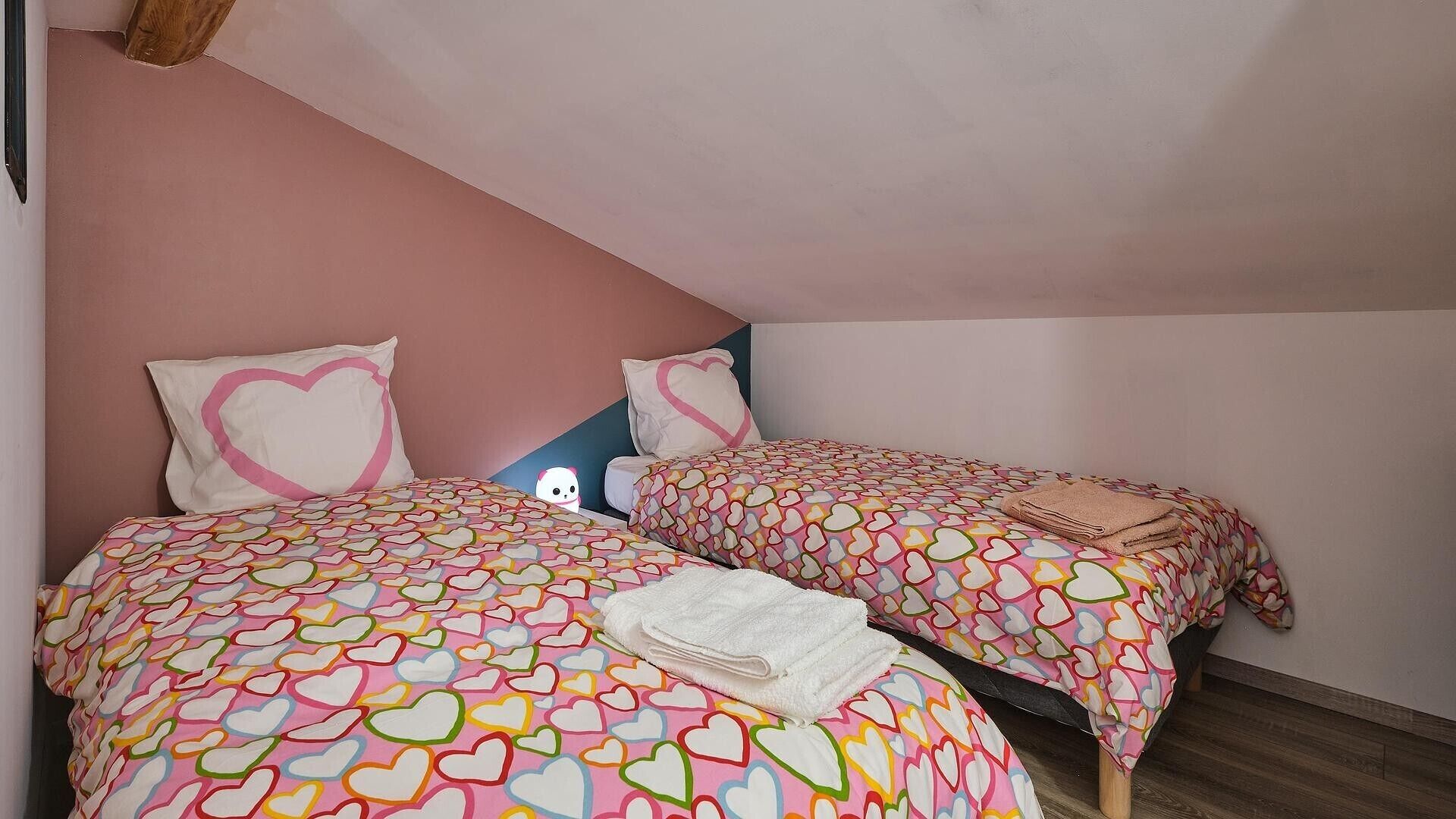 Un dormitorio con dos camas, cada una con ropa de cama estampada con motivos de corazón, una toalla doblada en una cama y una pared azul con una viga de madera.
