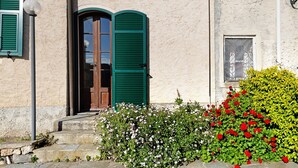 Un bâtiment avec une porte en bois, des volets verts et une petite fenêtre ornée d’une grille métallique.