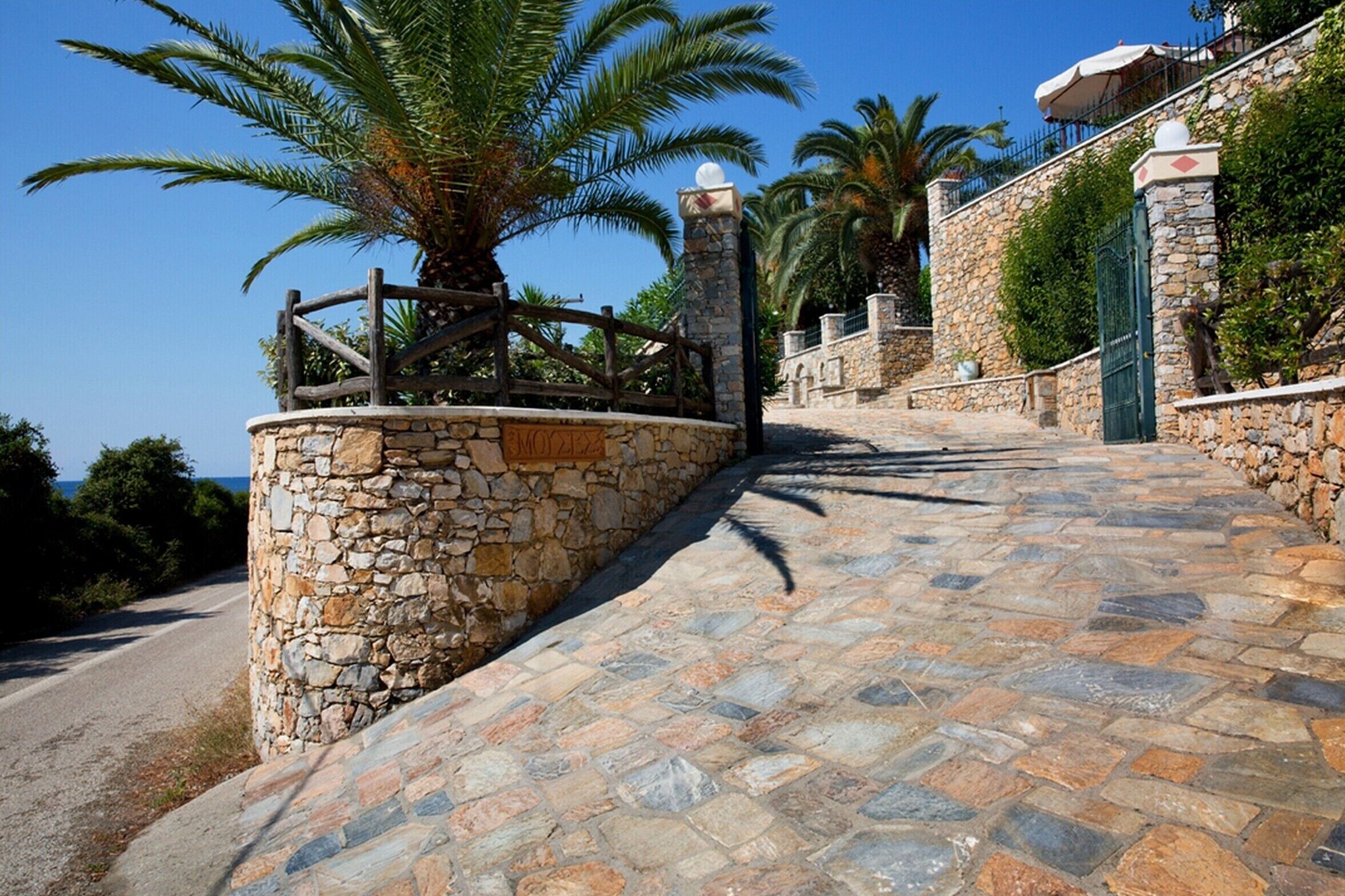 A stone pathway leading to a building with a stone wall and a palm tree.