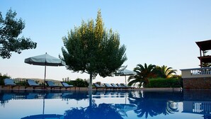 A swimming pool with lounge chairs and umbrellas, surrounded by trees and a building with a balcony. - Villa 'Beach  Alonissos For 6 People' with Shared Pool, Wi-Fi and Air Conditioning (Alonissos)
