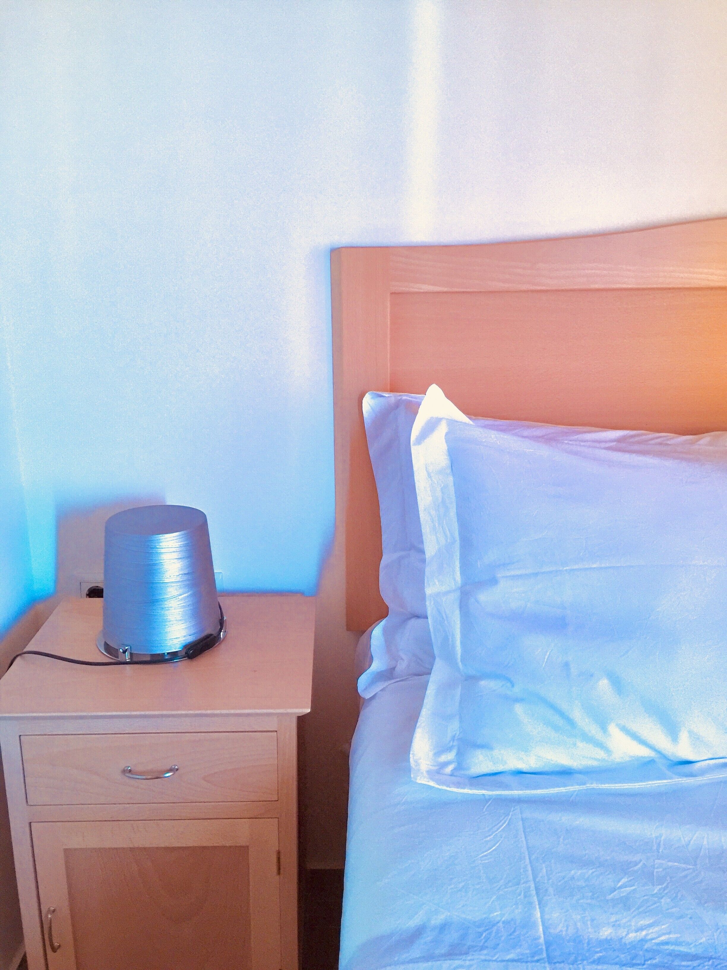 Un lit avec du linge de lit blanc, une tête de lit en bois et une table de chevet sur laquelle se trouve une bobine de fil.