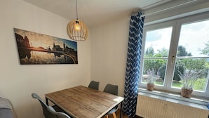 Un'area pranzo con tavolo e sedie in legno, un lampadario, una finestra con tende e un dipinto con vista sulla città.
