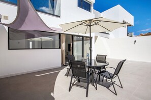 Une terrasse extérieure moderne avec une table en verre, des chaises noires, un parasol beige et un bâtiment blanc en arrière-plan.