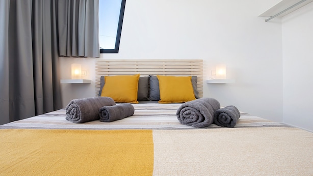 Een netjes opgemaakt bed met opgerolde handdoeken, een hoofdbord met twee wandlampen en een raam met gordijnen.