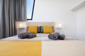 Un lit bien fait, avec des serviettes pliées, une tête de lit agrémentée de deux lampes fixées au mur, et une fenêtre avec des rideaux.