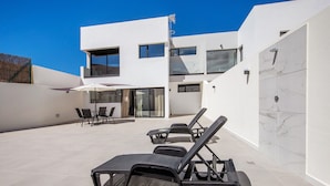 Een modern buitenverblijf met lounge fauteuils en een tafel, een wit gebouw met grote ramen, en een helderblauwe lucht.