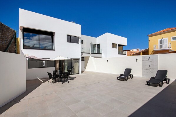 Un espace patio extérieur moderne, avec une table et des chaises pour les repas, des fauteuils de détente et un ciel d’un bleu limpide.