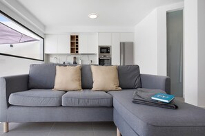 Un salon moderne avec un canapé gris, une petite table sur laquelle repose un livre, et une cuisine en arrière-plan.