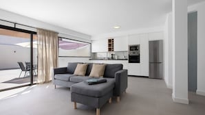 Een moderne woonkamer met een grijze modulaire bank, een eethoek met een tafel en stoelen, en een keuken met witte kasten en roestvrijstalen apparatuur.