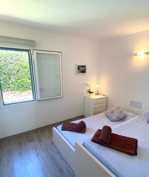 Une chambre à coucher avec un lit, une table de chevet, une petite table de nuit avec une lampe, une fenêtre et un petit tableau accroché au mur.