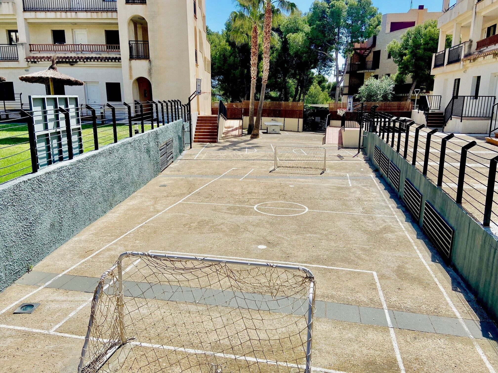 Una cancha de baloncesto exterior con aro, rodeada por una valla baja y adyacente a un edificio con balcones.