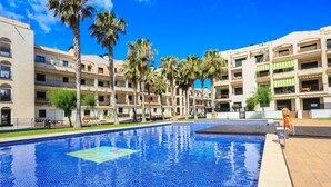 A swimming pool with tiled blue mosaic, surrounded by multi-story buildings with balconies and palm trees.