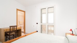Un dormitorio con cama, escritorio, silla, mesita de noche con lámpara y puerta que da a un balcón.