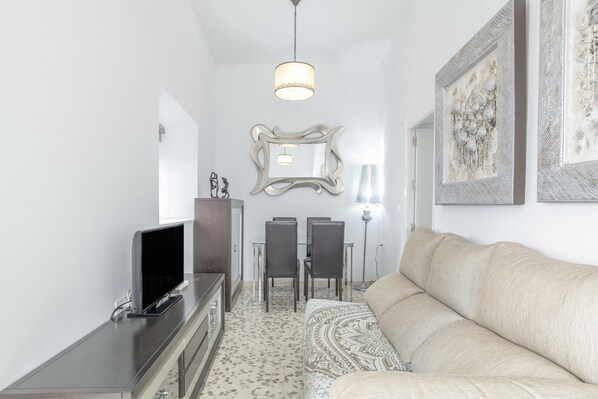 A living room with a sofa, a TV on a stand, a dining table with chairs, and a mirror on the wall.