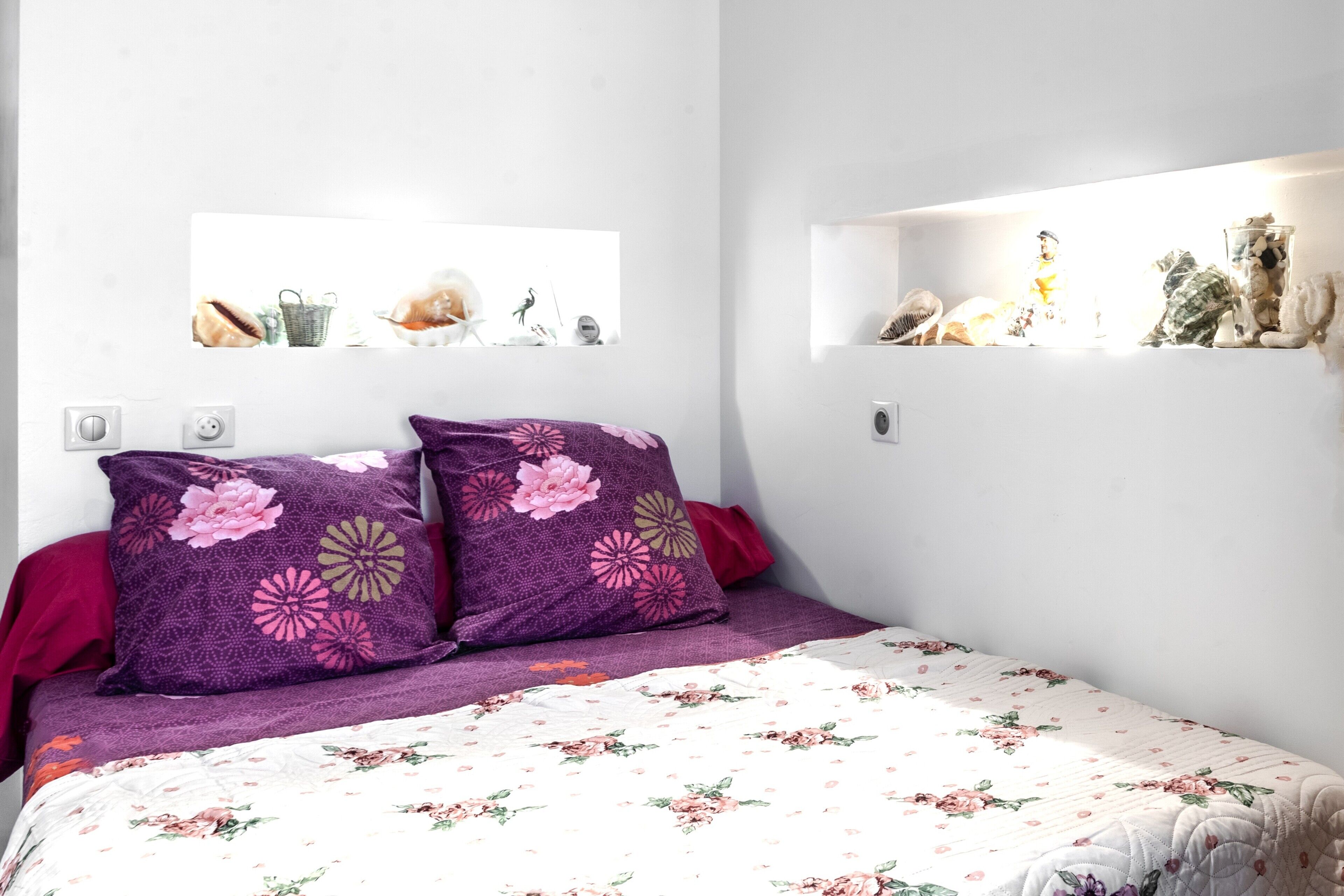 A bedroom with a floral bedspread, purple pillows, and wall shelves with decorative items.