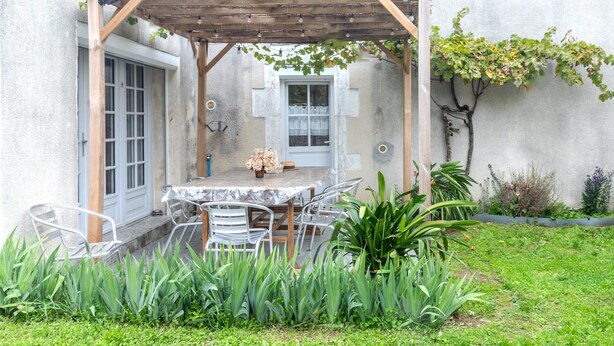 A covered patio with a table and chairs, a pergola, and a garden with plants and a wall.