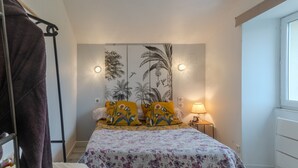 A bedroom with a bed, a nightstand, a lamp, a wall with a tropical mural, and a window. - Holiday Home 'La Maison D'Éloi' with Shared Pool, Private Terrace & Wi-Fi (Montignac-Charente)