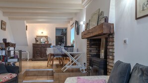 A living room with a fireplace, a sofa, a dining area with chairs, and a kitchen in the background. - Holiday Home 'La Maison D'Éloi' with Shared Pool, Private Terrace & Wi-Fi (Montignac-Charente)