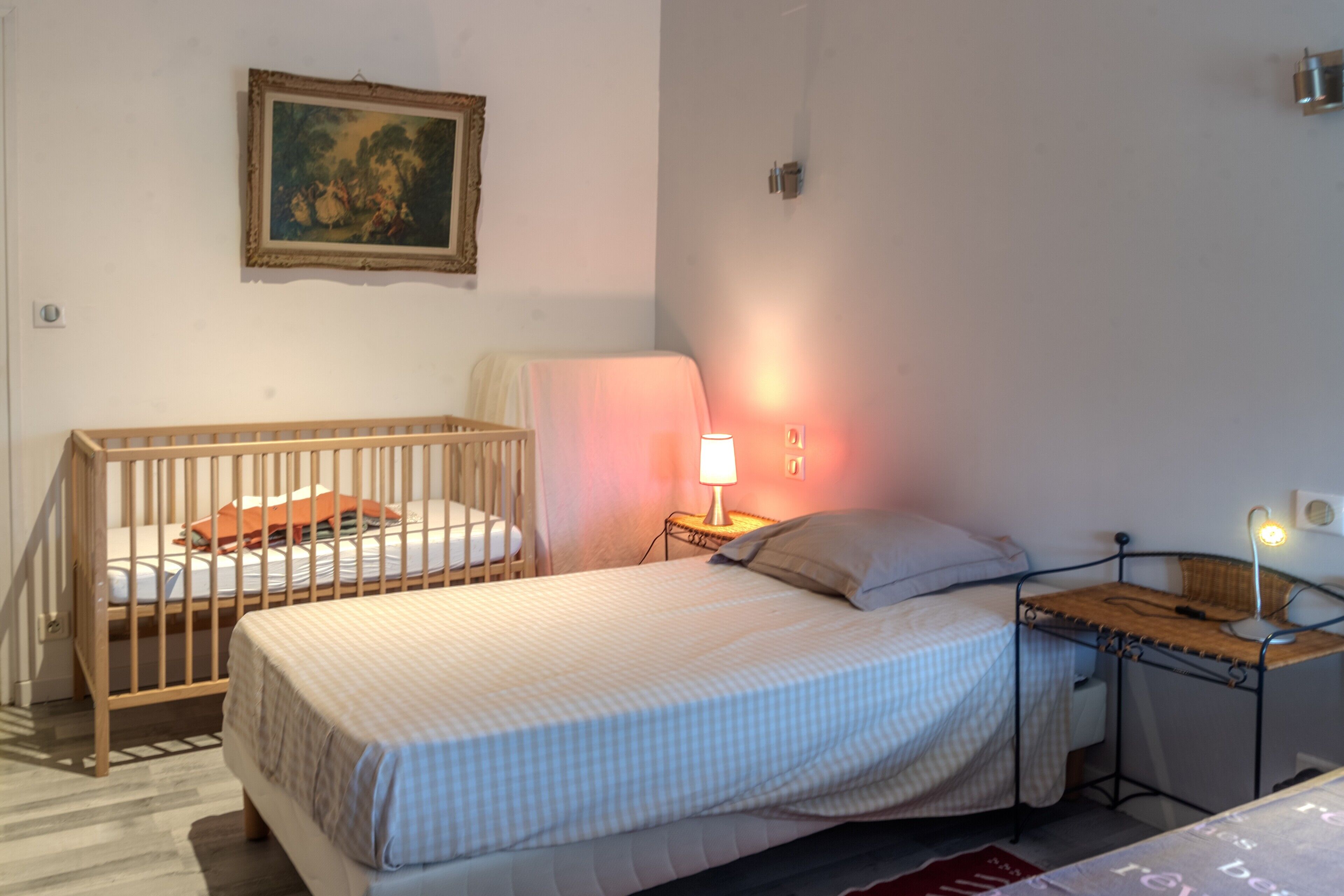 A bedroom with a crib, a bed, a nightstand with a lamp, and a wall-mounted picture.
