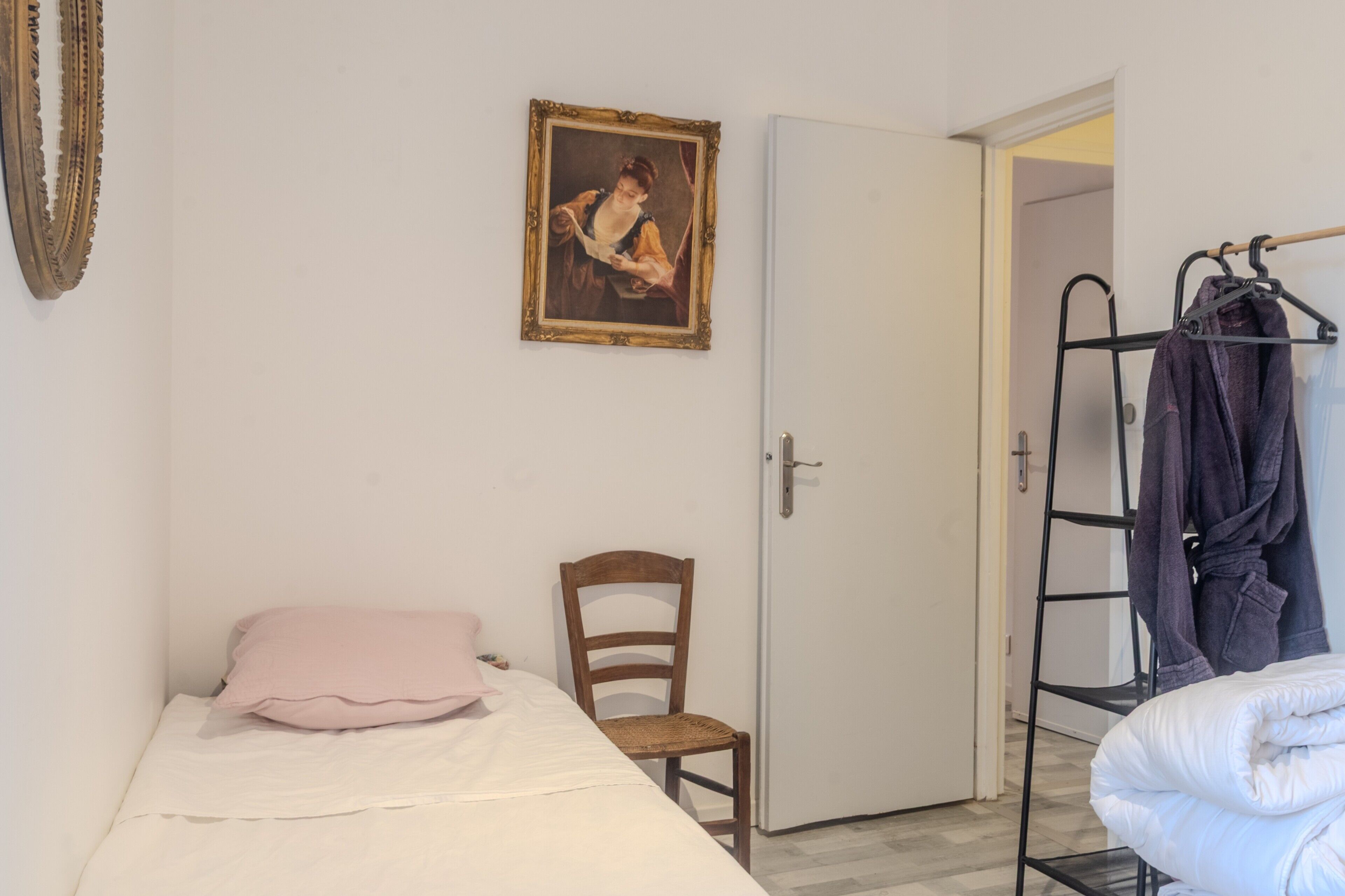 A room with a bed, a chair, a door, a coat rack, and a framed picture on the wall.
