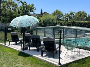 Ein überdachter Außenbereich mit Stühlen und Sonnenschirm, ein Schwimmbecken und ein umzäunter Garten.