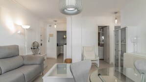 A modern living room with a sofa, a glass-top coffee table, a chair, and a dining table. There is a kitchen in the background with cabinets and a refrigerator. - Casa Ricardo - Vacation Home Near the Beach in Barbate (Barbate)