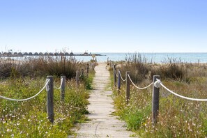 Ein Holzsteg führt zu einem Strand, im Hintergrund ist ein Steg zu sehen.