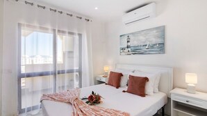 A hotel room with a bed, a nightstand, a lamp, a painting, and a view of buildings through sheer curtains. - Apartment 'Penthouse Paradise - Praia Da Rocha' with Sea View, Wi-Fi and Air Conditioning (Portimão)