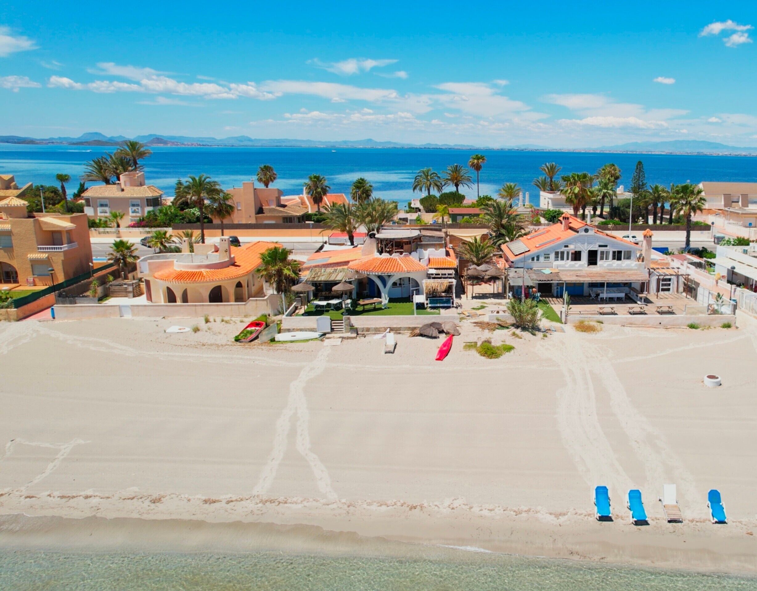 An aerial view of a coastal area with buildings, palm trees, and a beach with lounge chairs.