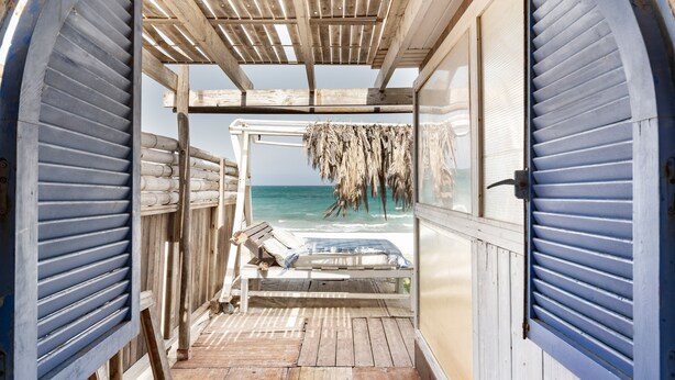 Une terrasse en bois menant à un point de vue sur la plage, avec un fauteuil de relaxation et une structure dotée d’un toit de chaume.
