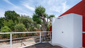 Un balcón con piso de madera, barandal blanco y vista a árboles y un edificio rojo.