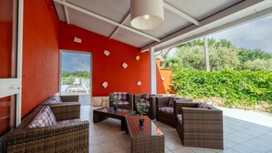 Un espacio de estar al aire libre con techo, mobiliario de mimbre y una mesa de centro de vidrio, una pared roja y vistas a zonas verdes.
