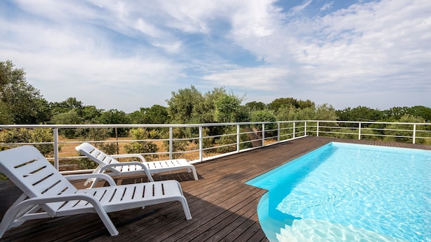 Una terraza de madera con piscina, sillas de descanso blancas y vistas a zonas verdes.