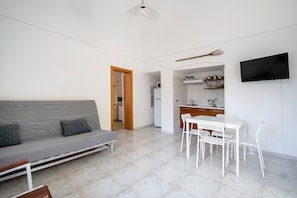 Un salon moderne comprenant un canapé, une table à manger avec des chaises, un coin cuisine et un téléviseur à écran plat fixé au mur.