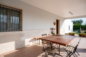 Un patio exterior con una mesa y sillas de madera, una ventana con rejas y plantas en macetas.