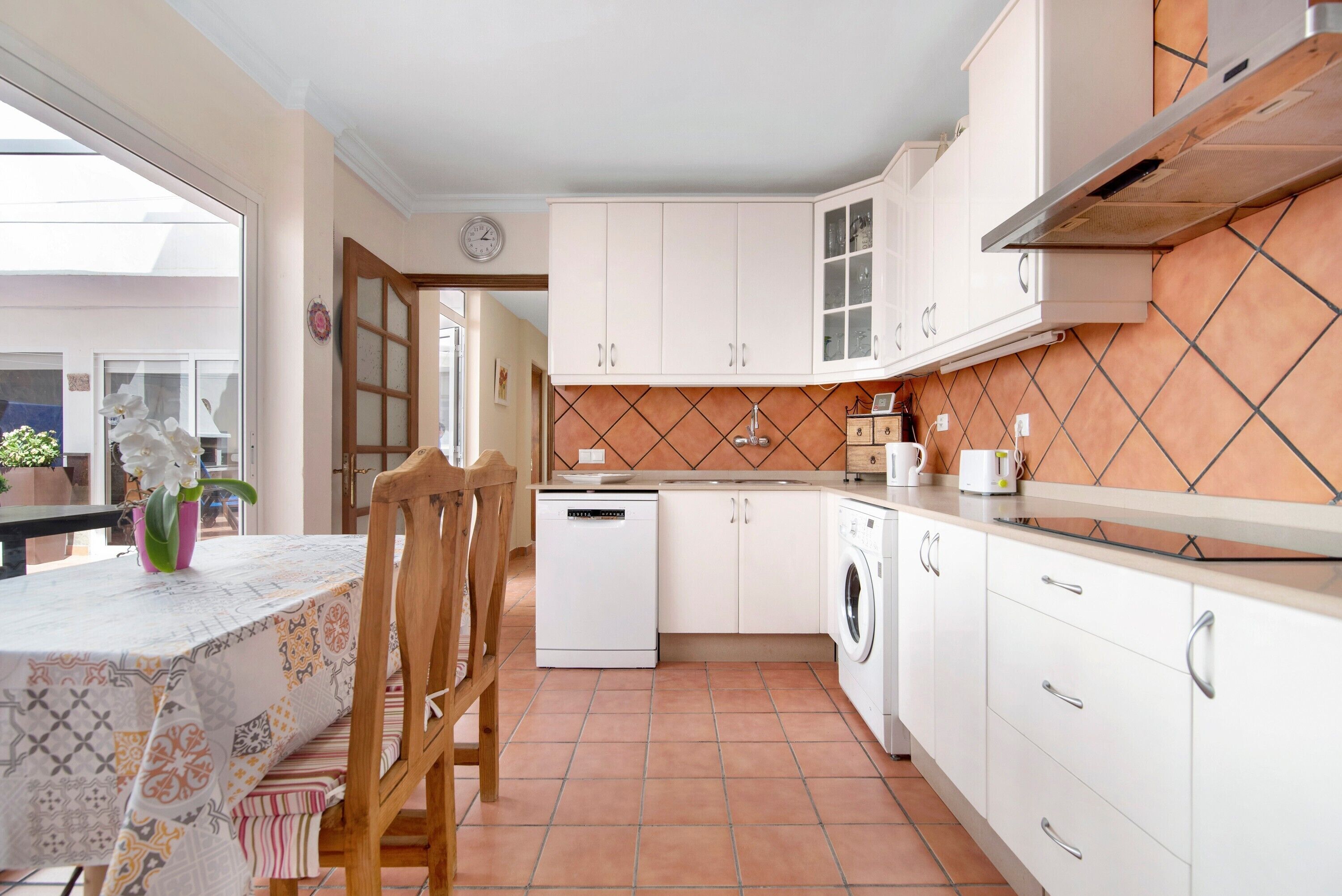 Una cocina con armarios blancos, lavavajillas, lavadora y un comedor con mesa y sillas.