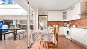 Une cuisine avec des meubles blancs, un lave-linge et une table à manger recouverte d’une nappe à motifs.