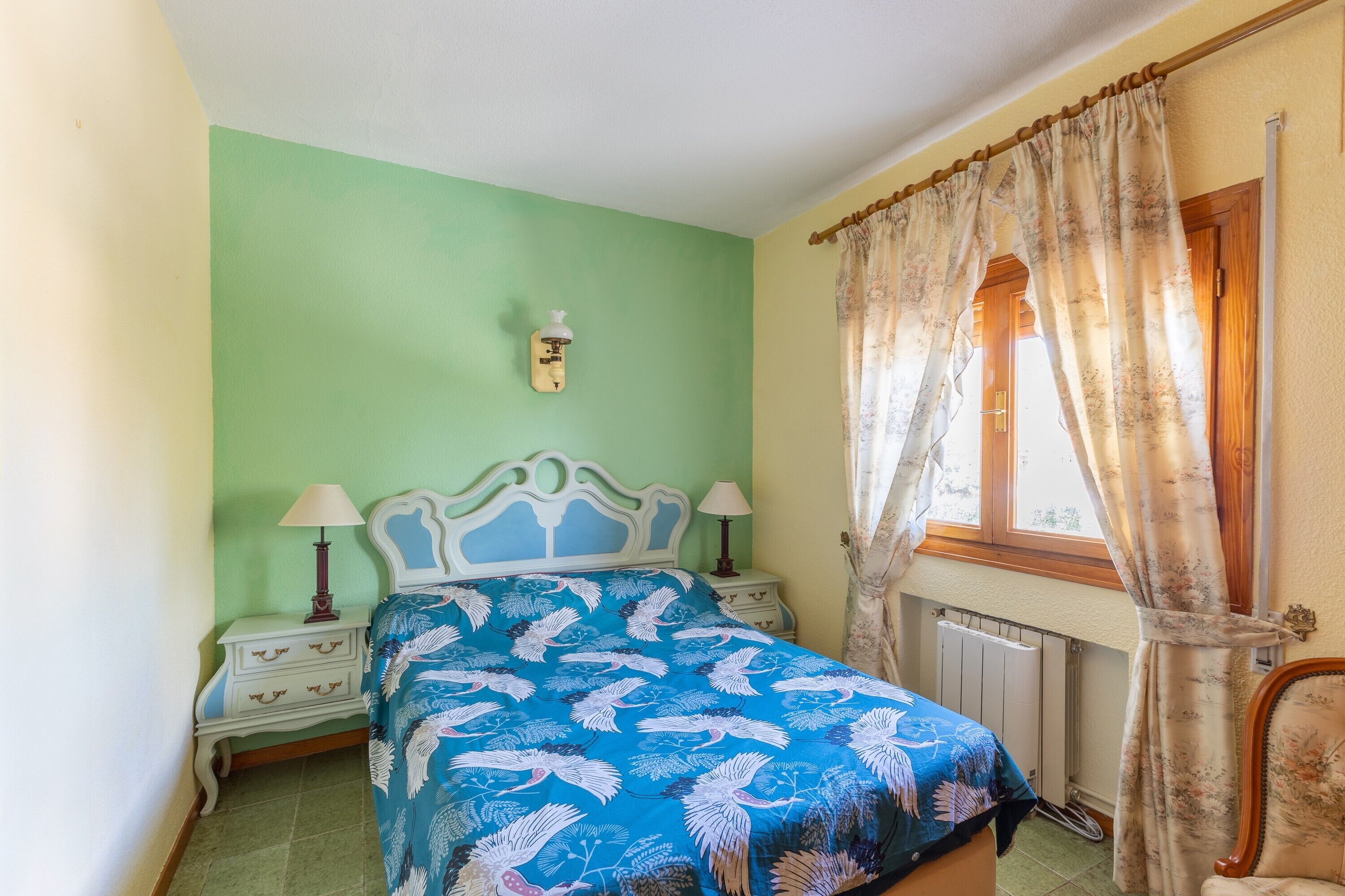 Un dormitorio con cama, mesitas de noche, un radiador y una ventana con cortinas.