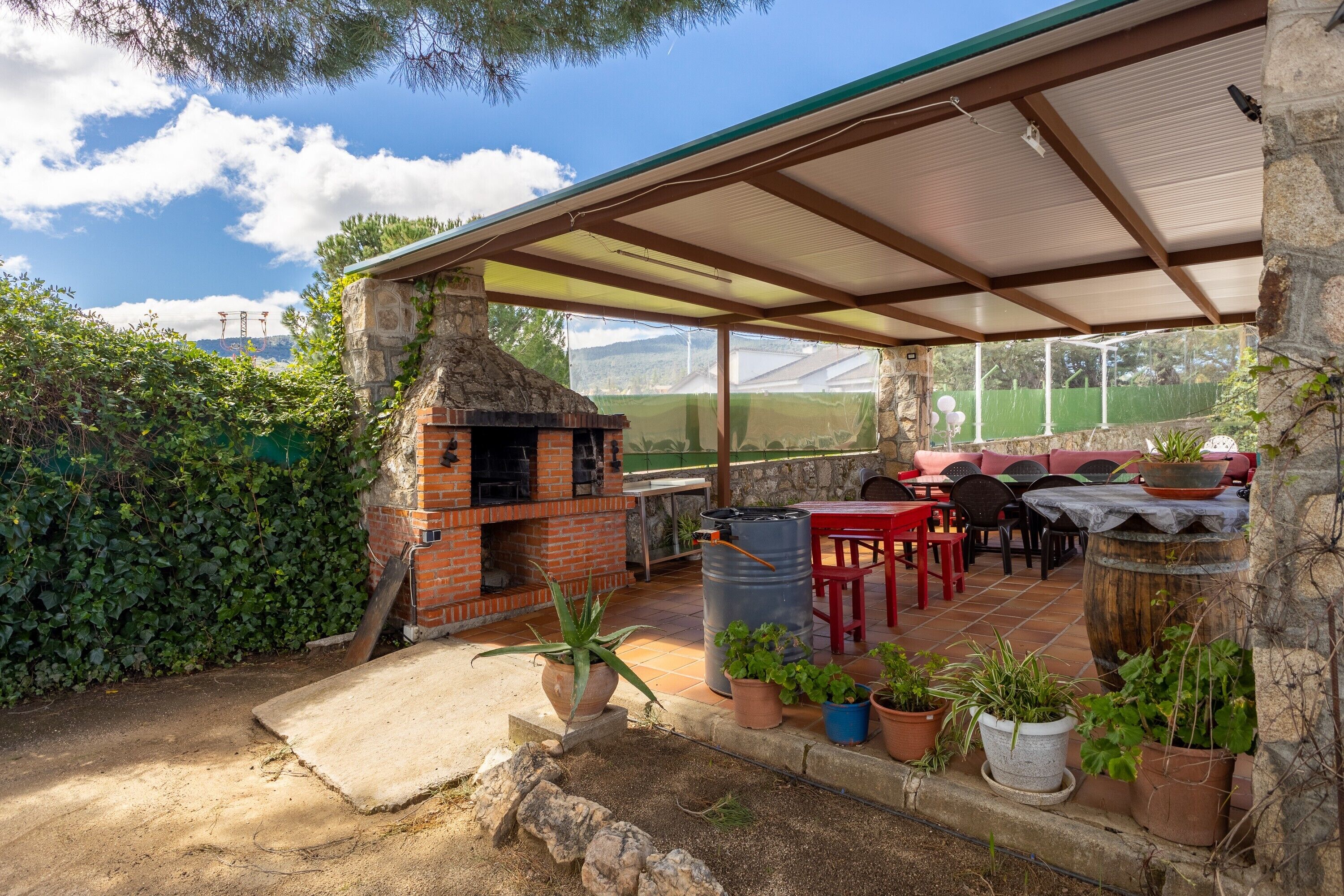 Een terras buiten met een bakstenen barbecue, potplanten en een overdekte zithoek.