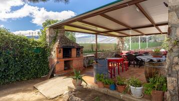 Een terras buiten met een bakstenen barbecue, potplanten en een overdekte zithoek.