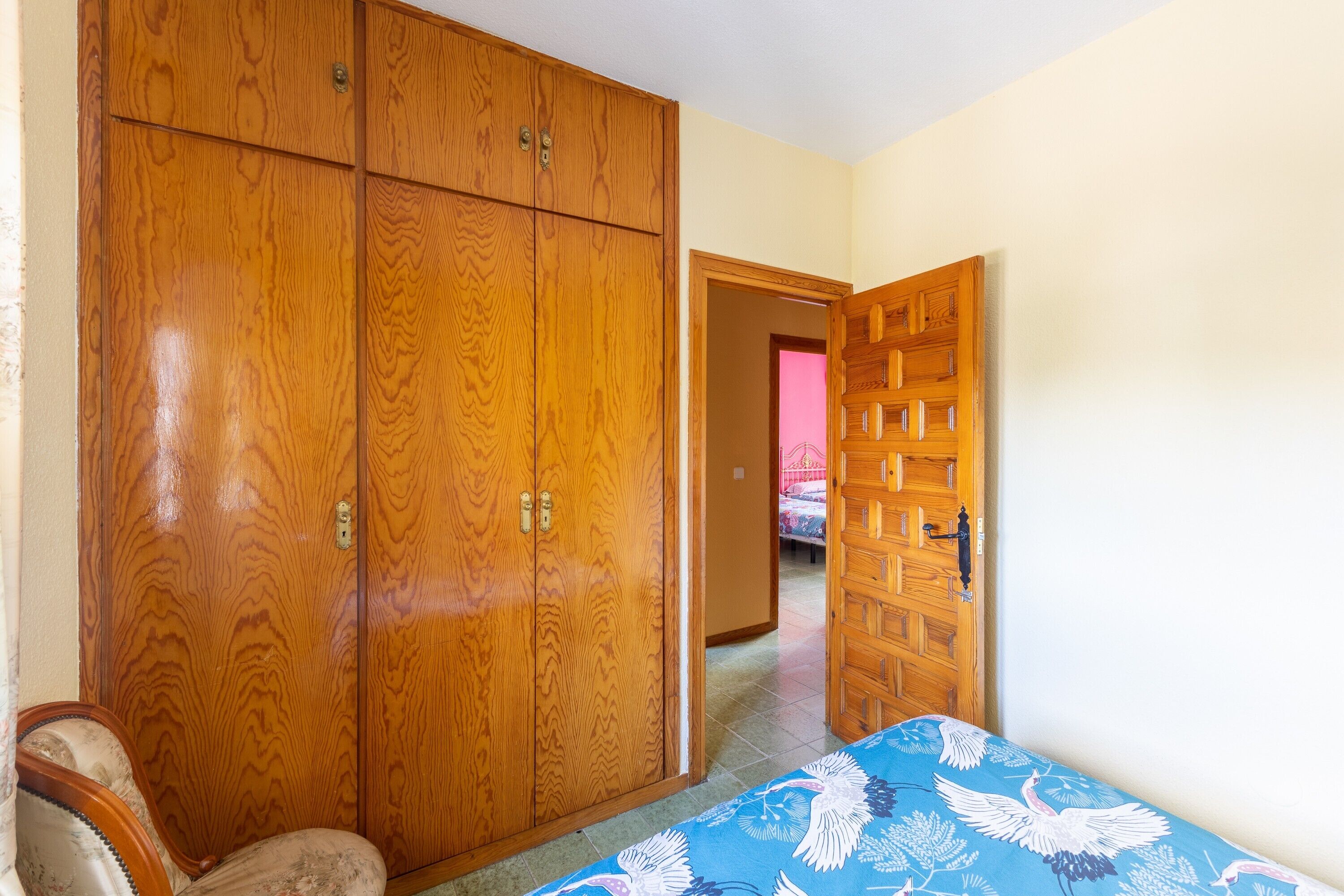 Un dormitorio con un armario de madera, una cama con colcha estampada y una puerta que da a otra habitación.