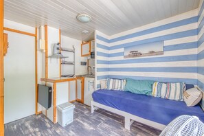 Une chambre avec un lit, une petite kitchenette, un ventilateur blanc et un tableau accroché au mur.