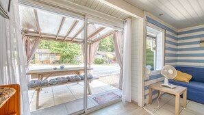 A room with a sliding glass door leading to an outdoor area, a small table with a fan, and a blue-striped wall. - Apartment 'Chalet B Village Vacances' with Shared Pool, Private Terrace and Wi-Fi (Gujan-Mestras)